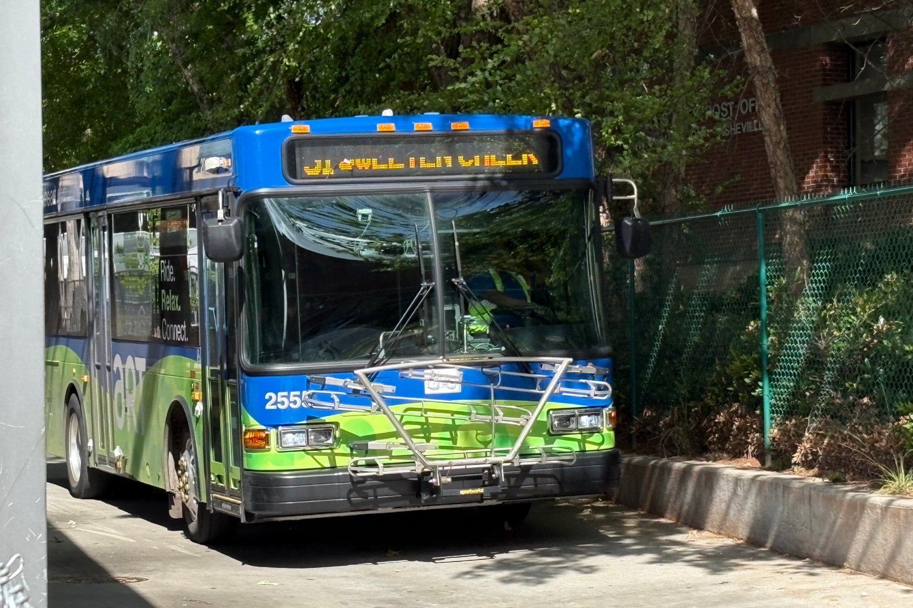 Front view of a blue and green city bus with a bike rack on the bumper and an amber destination sign above the windshield, parked on a street.