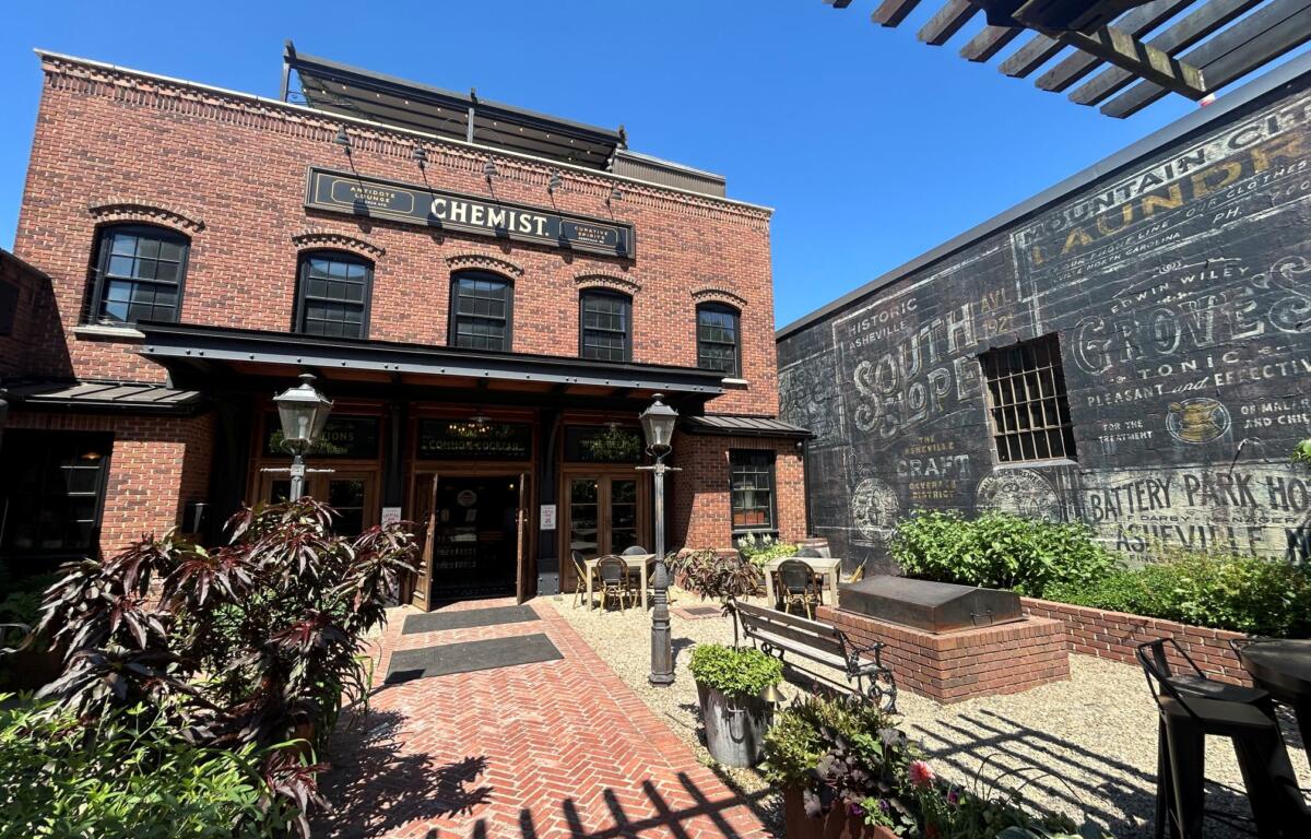 Brick storefront with a sign reading 'CHEMIST' over the doorway, outdoor seating, and string lights in a sunny courtyard. A mural-covered wall is to the right.