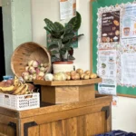 Wooden market stand with baskets of onions and potatoes, bananas, and a potted plant beside a bulletin board of posters.