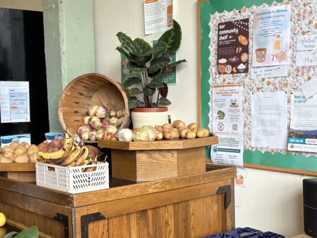 Wooden market stand with baskets of onions and potatoes, bananas, and a potted plant beside a bulletin board of posters.