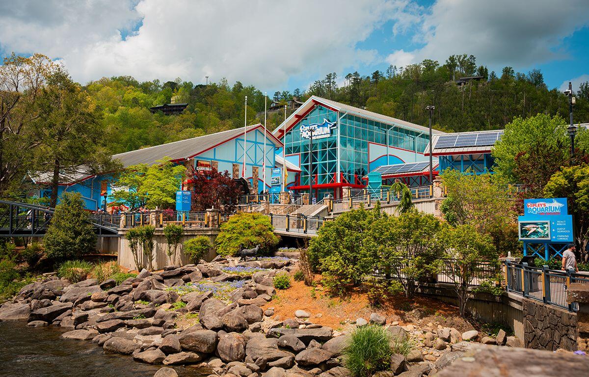 Exterior view of Ripley's Aquarium with blue and red-framed glass facade, set against a hillside with trees and a rocky waterfront in the foreground