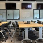 Conference room with long wooden tables arranged in a U-shape, black chairs, phones, and wall-mounted TVs above the glass wall panels.