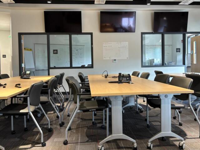 Conference room with long wooden tables arranged in a U-shape, black chairs, phones, and wall-mounted TVs above the glass wall panels.