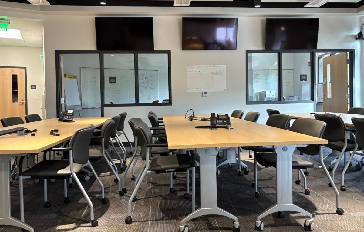 Conference room with long wooden tables arranged in a U-shape, black chairs, phones, and wall-mounted TVs above the glass wall panels.