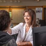 Optometrist in a white coat smiling at a patient across a desk with a computer monitor in an eyeglasses clinic setting.