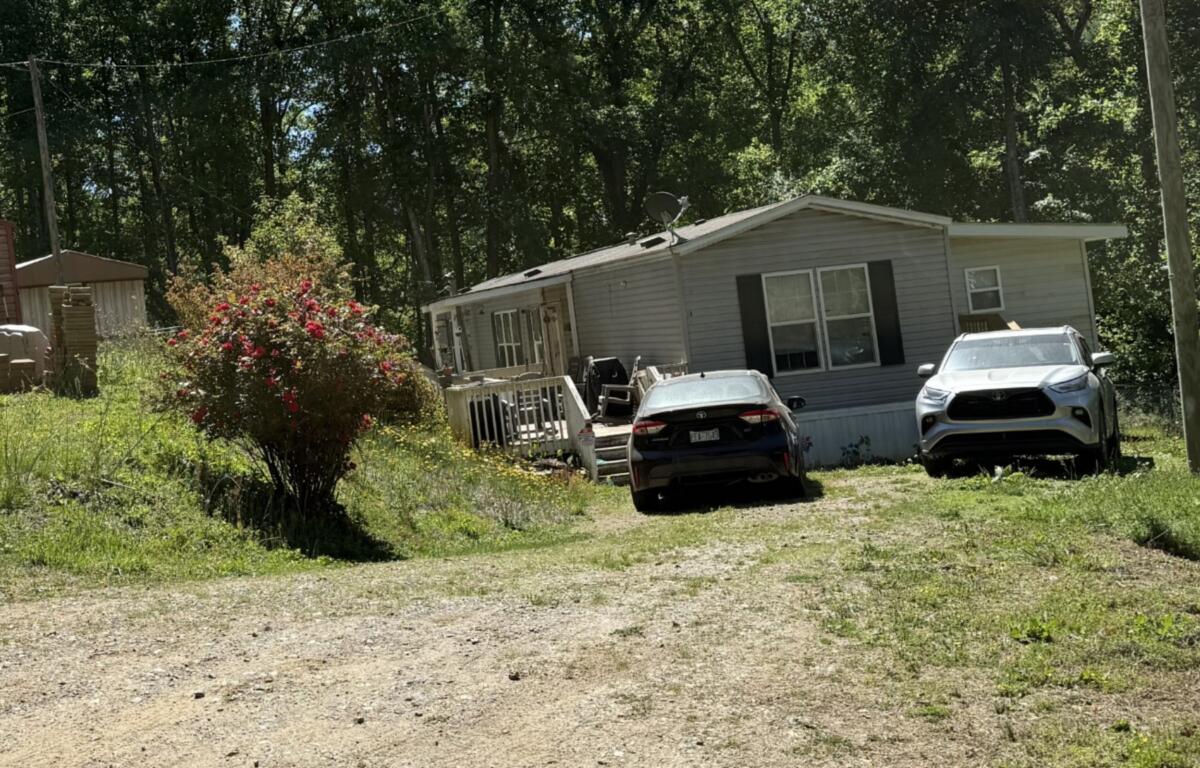 A small single-story mobile home with two cars parked in the yard, dirt driveway, and surrounding trees and bushes.