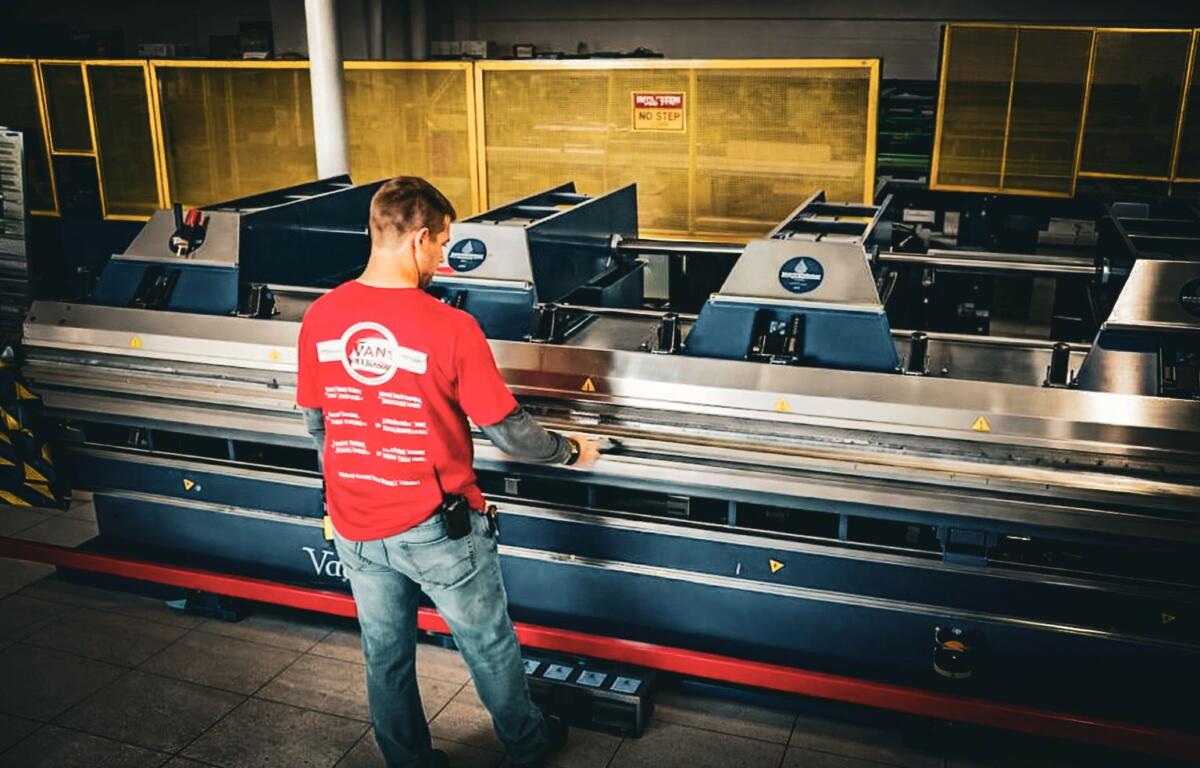 Factory worker in a red shirt operating a large industrial machine with metal rollers in a dimly lit workshop.