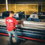 Factory worker in a red shirt operating a large industrial machine with metal rollers in a dimly lit workshop.