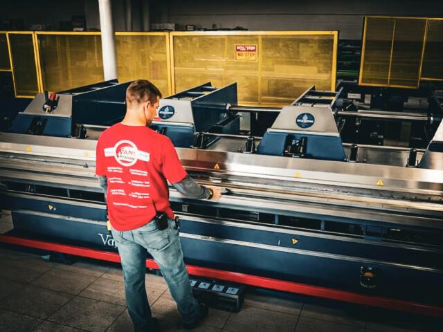 Factory worker in a red shirt operating a large industrial machine with metal rollers in a dimly lit workshop.