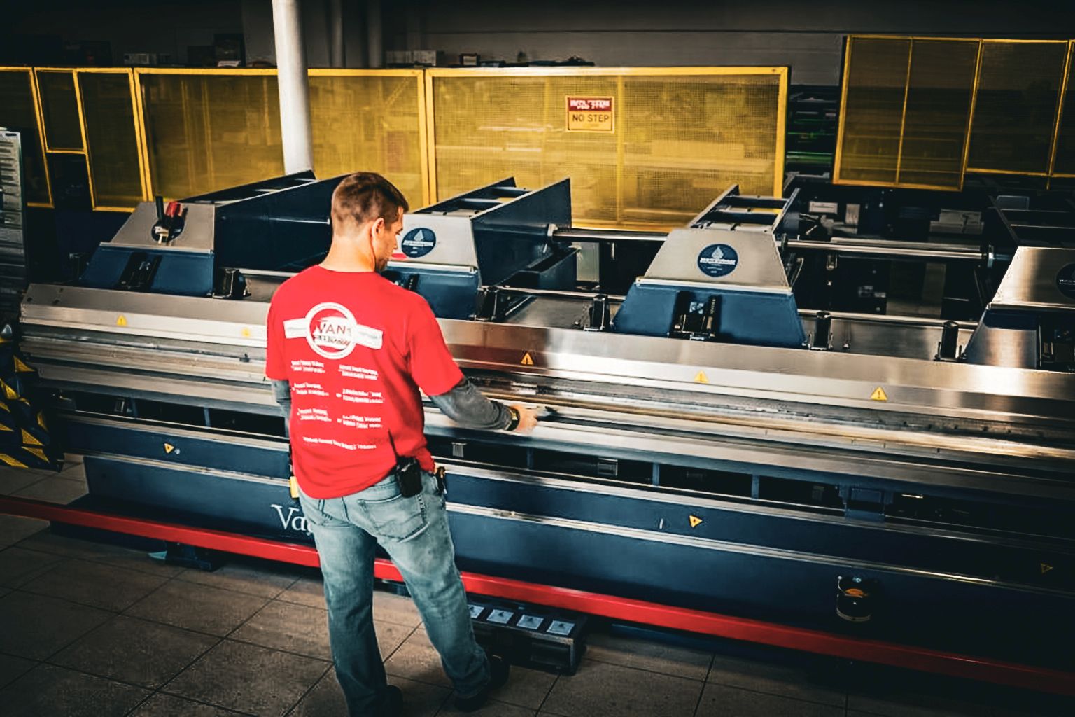Factory worker in a red shirt operating a large industrial machine with metal rollers in a dimly lit workshop.