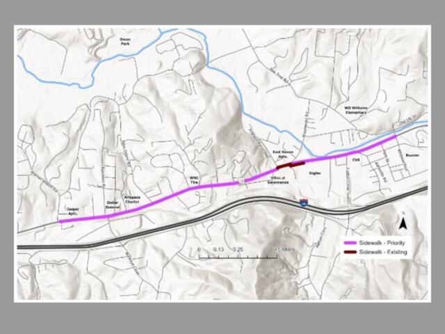 Map of a town showing a proposed sidewalk route in purple (priority) along a highway, with an existing sidewalk in maroon and blue water features nearby.