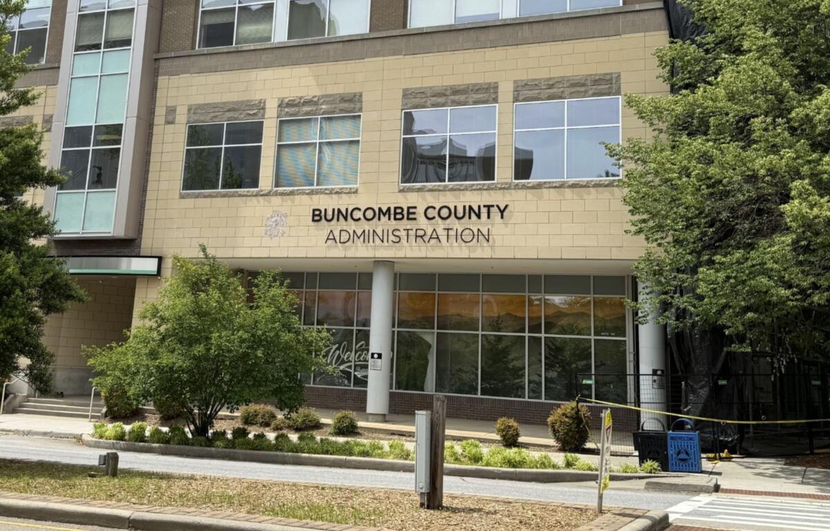 Front of Buncombe County Administration building with a beige brick facade, tall windows, and surrounding trees; sign reads 'BUNCOMBE COUNTY ADMINISTRATION' above the entrance