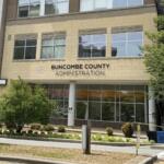 Front of Buncombe County Administration building with a beige brick facade, tall windows, and surrounding trees; sign reads 'BUNCOMBE COUNTY ADMINISTRATION' above the entrance