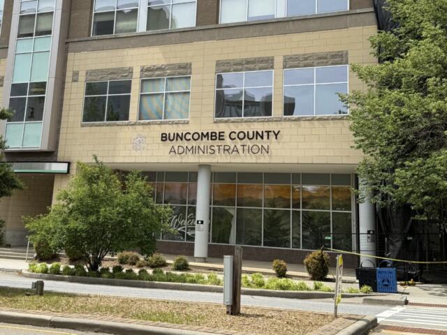 Front of Buncombe County Administration building with a beige brick facade, tall windows, and surrounding trees; sign reads 'BUNCOMBE COUNTY ADMINISTRATION' above the entrance