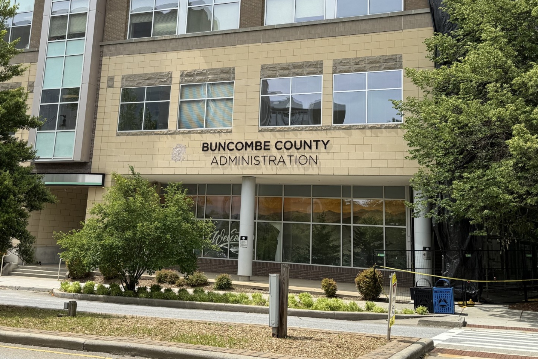 Front of Buncombe County Administration building with a beige brick facade, tall windows, and surrounding trees; sign reads 'BUNCOMBE COUNTY ADMINISTRATION' above the entrance