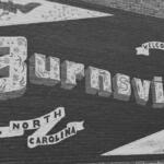 Brick-wall mural featuring ornamental text 'Burlington' with a 'Welcome' banner and ribbons reading 'North Carolina'—a city welcome sign.
