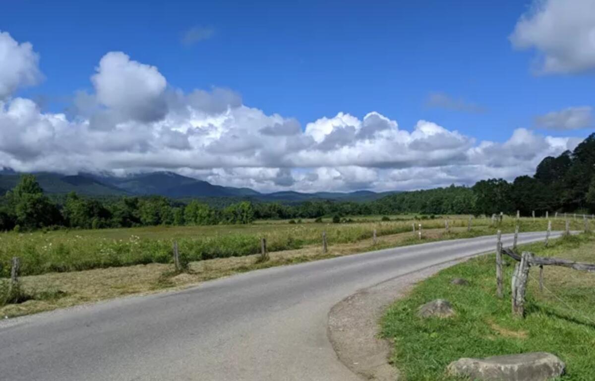 Paved country road with a wooden fence, green fields, and distant hills under a bright blue sky and white clouds.