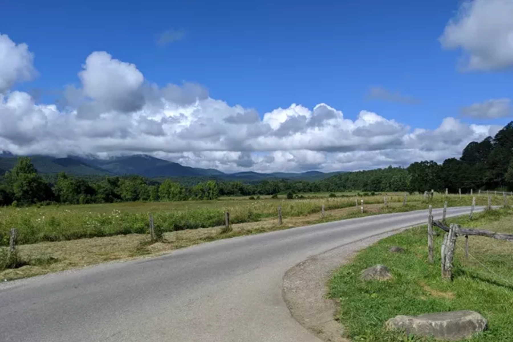 Paved country road with a wooden fence, green fields, and distant hills under a bright blue sky and white clouds.