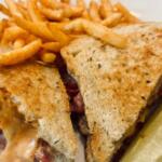 Grilled sandwich cut into triangles with melted cheese and meat, served with French fries and a pickle spear on a plate.