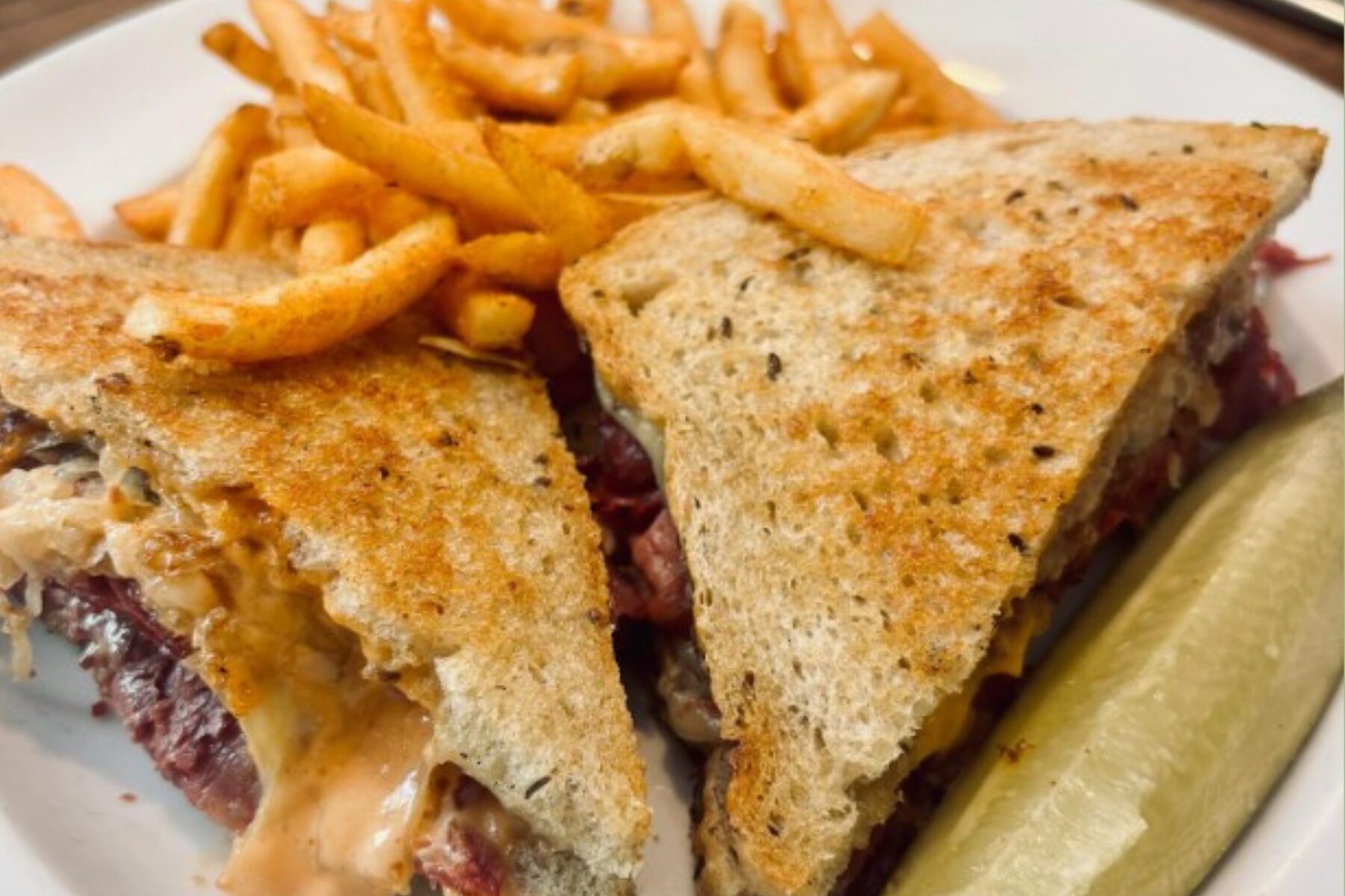 Grilled sandwich cut into triangles with melted cheese and meat, served with French fries and a pickle spear on a plate.