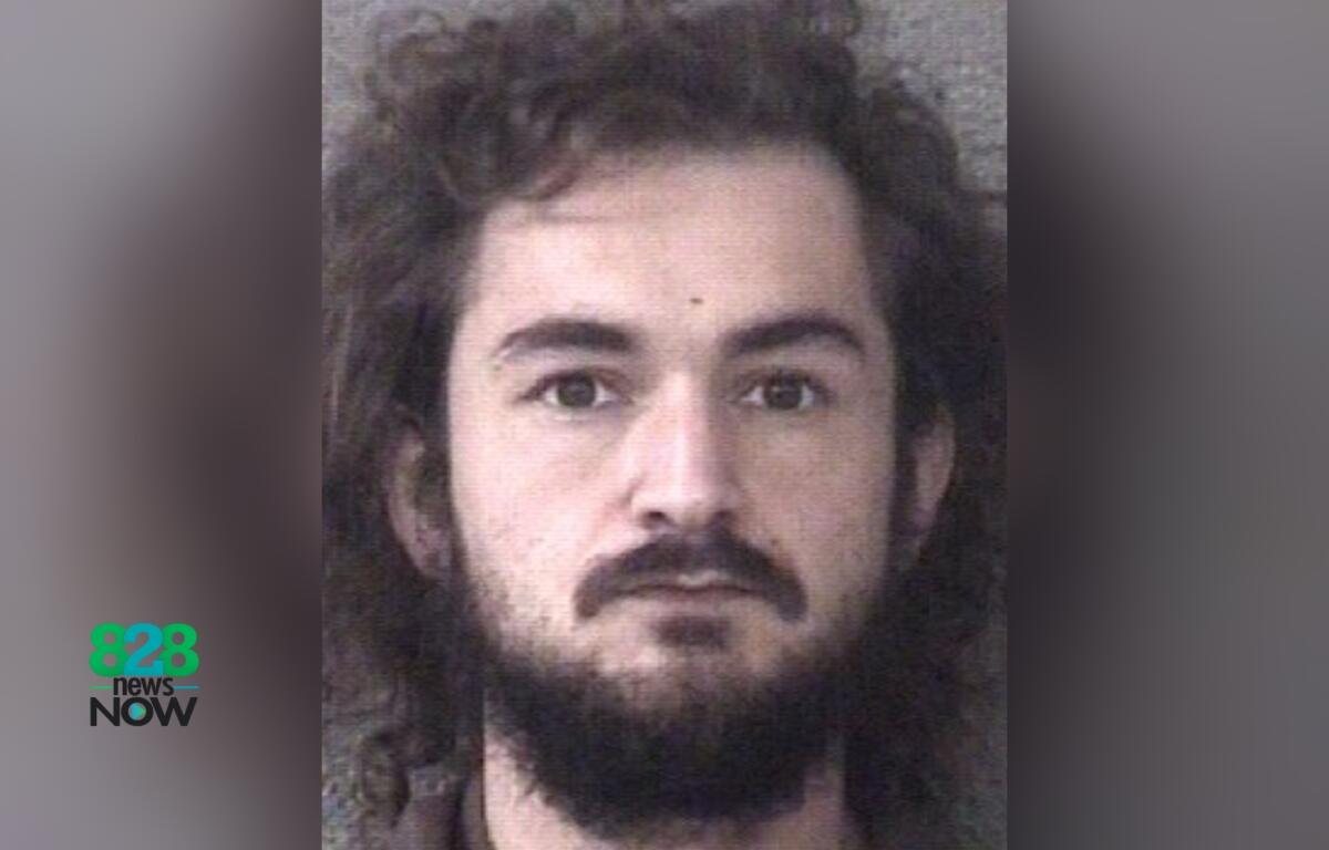 Mugshot-style head-and-shoulders photo of a man with long dark hair and a full beard, neutral backdrop, 828 News Now logo in the bottom left.