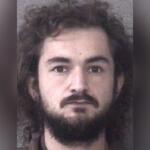 Mugshot-style head-and-shoulders photo of a man with long dark hair and a full beard, neutral backdrop, 828 News Now logo in the bottom left.