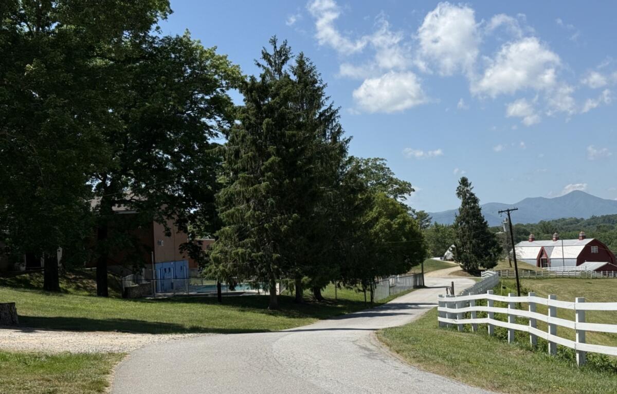 Rural road curving by trees and a white fence, with a red barn and blue sky in the distance. Mountains on the horizon.