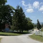Rural road curving by trees and a white fence, with a red barn and blue sky in the distance. Mountains on the horizon.