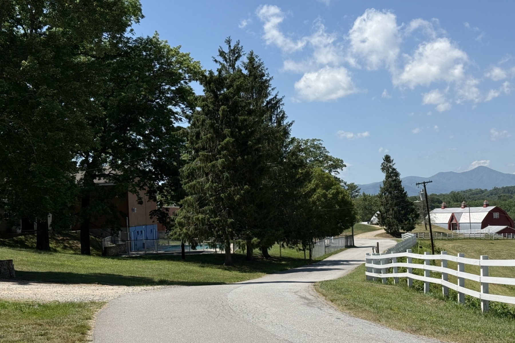 Rural road curving by trees and a white fence, with a red barn and blue sky in the distance. Mountains on the horizon.