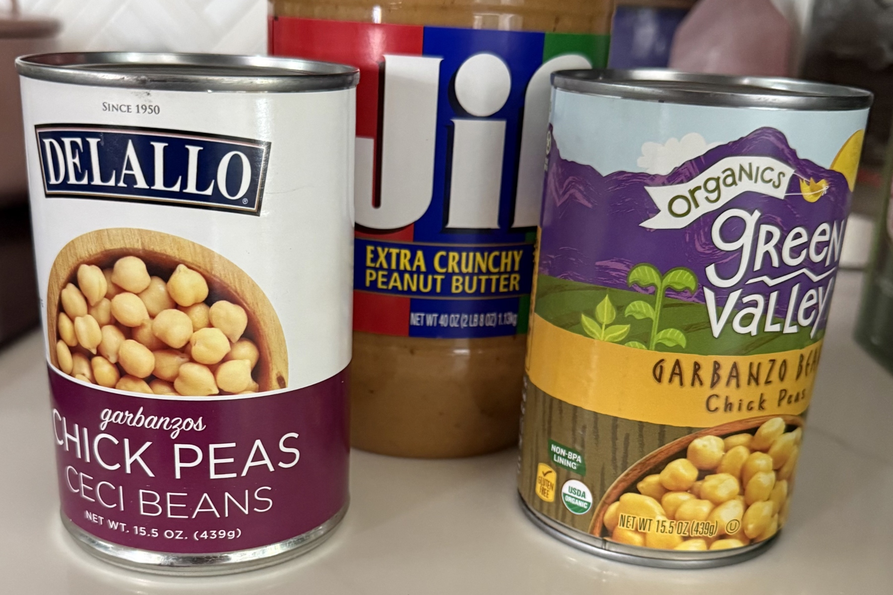Three canned pantry items: Delallo chickpeas (garbanzo beans) on the left, Jif Extra Crunchy Peanut Butter in the center, and Green Valley Organics garbanzo beans on the right.