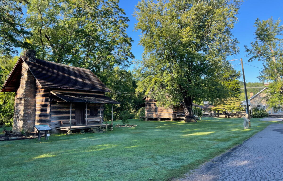 Rustic log cabin with a stone chimney beneath green trees in a sunny park next to a cobblestone path and grassy lawn.