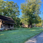 Rustic log cabin with a stone chimney beneath green trees in a sunny park next to a cobblestone path and grassy lawn.