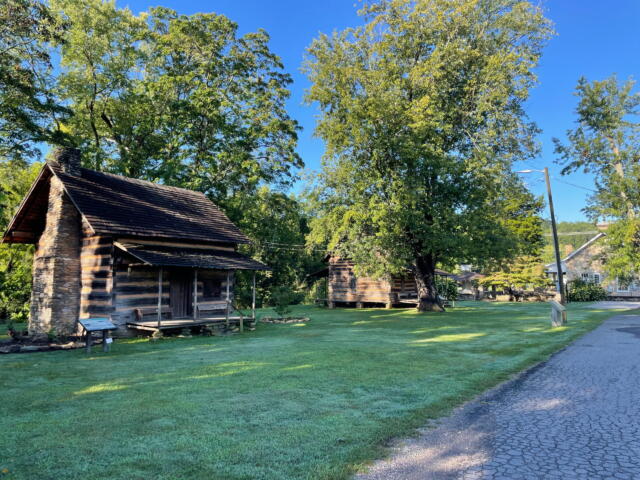 Rustic log cabin with a stone chimney beneath green trees in a sunny park next to a cobblestone path and grassy lawn.