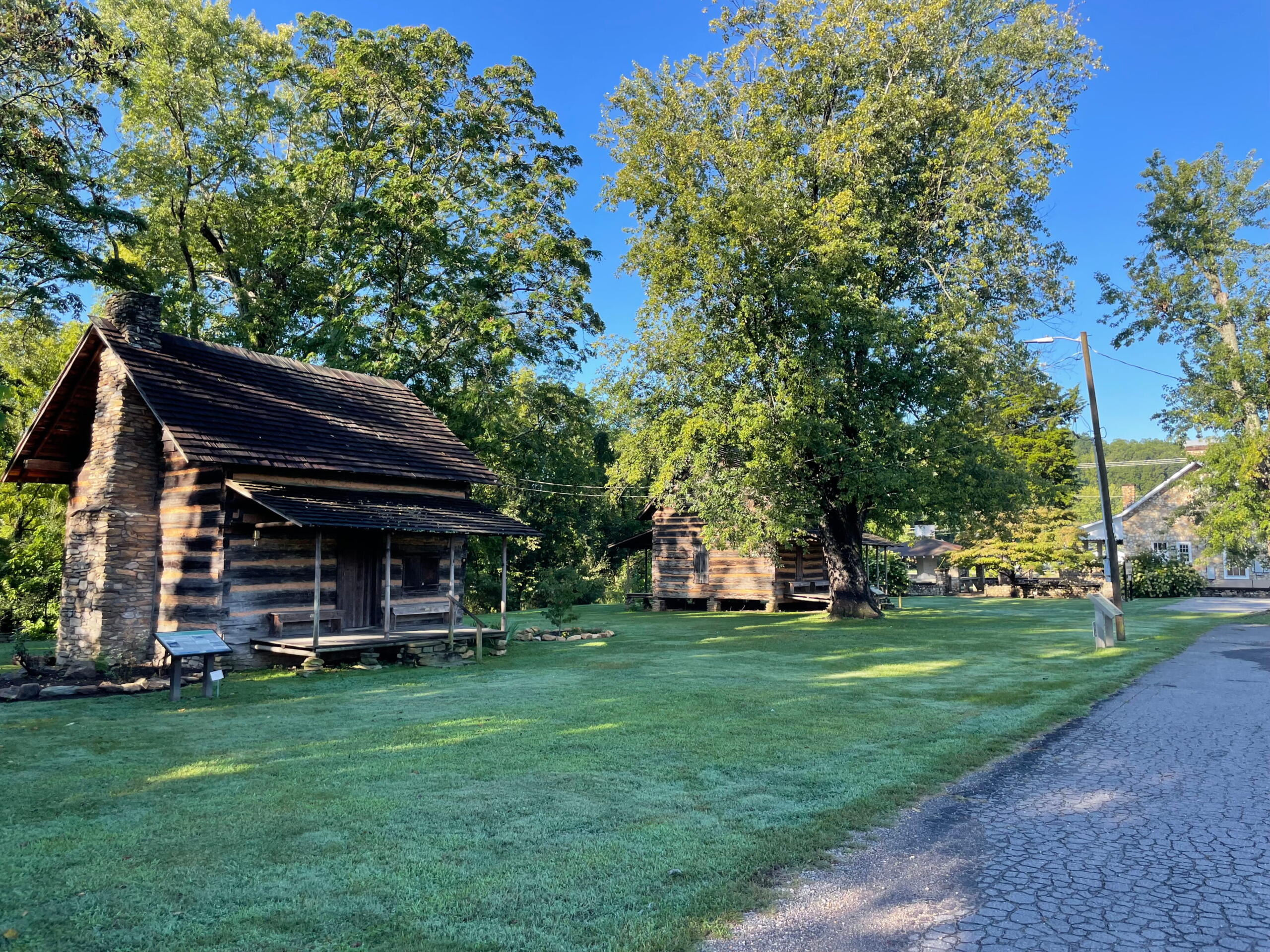 Rustic log cabin with a stone chimney beneath green trees in a sunny park next to a cobblestone path and grassy lawn.