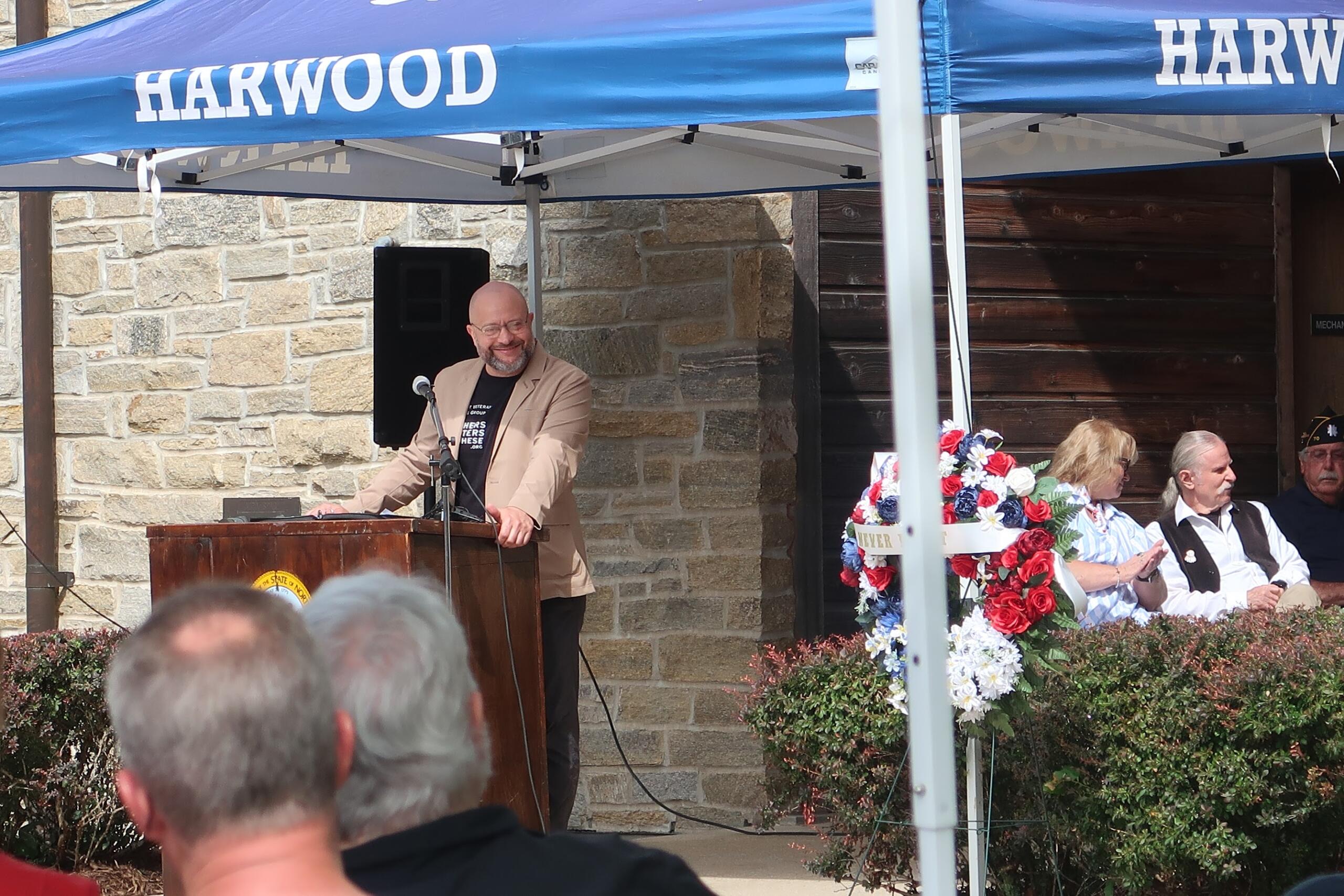 Man in a tan blazer speaks at a wooden podium under a blue canopy labeled HARWOOD, with a floral arrangement nearby and audience seated behind him.