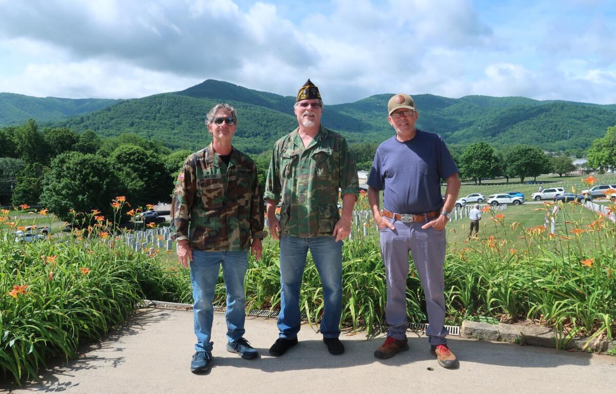 Three men stand side by side in a garden with orange lilies, mountains in the background.