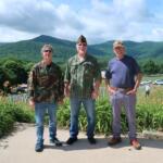 Three men stand side by side in a garden with orange lilies, mountains in the background.