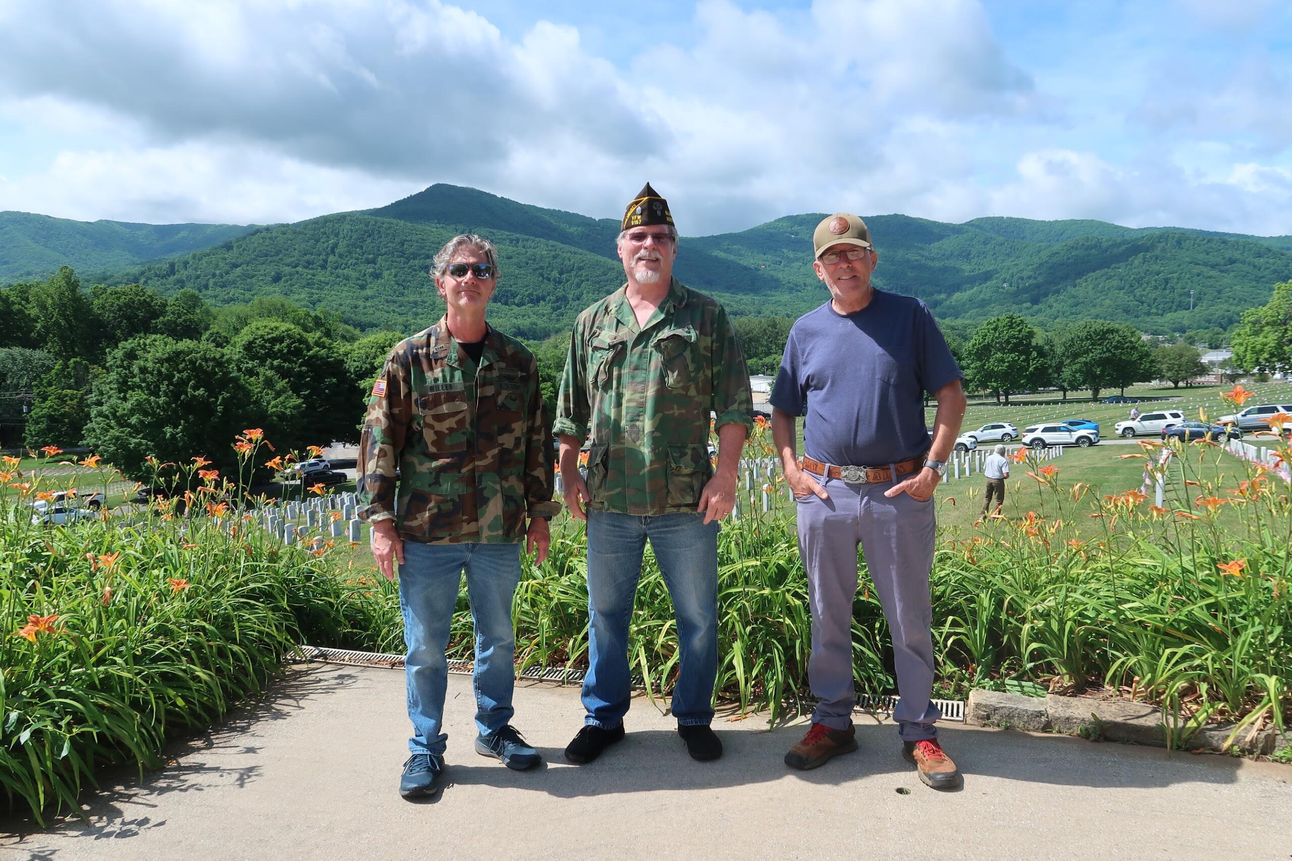 Three men stand side by side in a garden with orange lilies, mountains in the background.