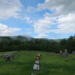 Graveyard with rows of headstones on a grassy slope, trees in the background under a partly cloudy blue sky.
