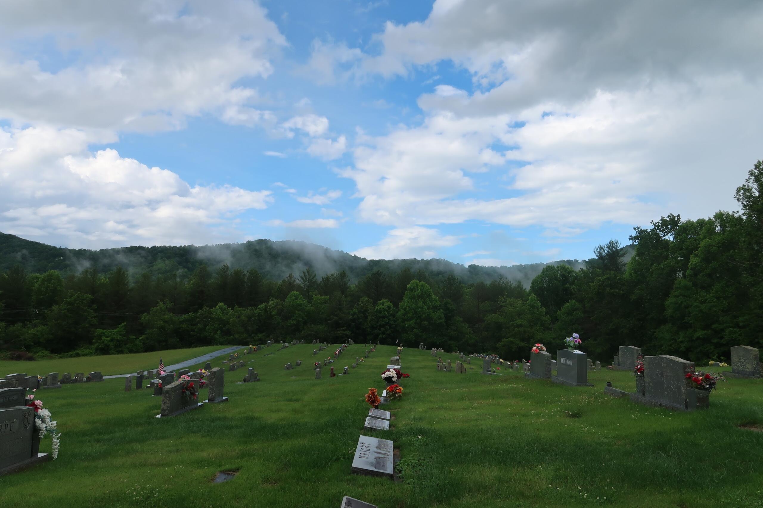 Graveyard with rows of headstones on a grassy slope, trees in the background under a partly cloudy blue sky.