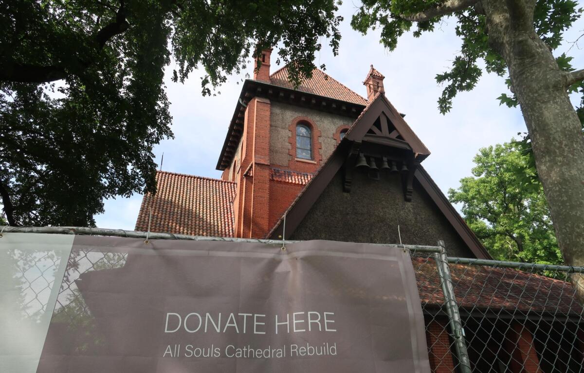 Brick church with red-tiled roof visible through surrounding trees; foreground banner reads “Donate Here” for All Souls Cathedral rebuild.