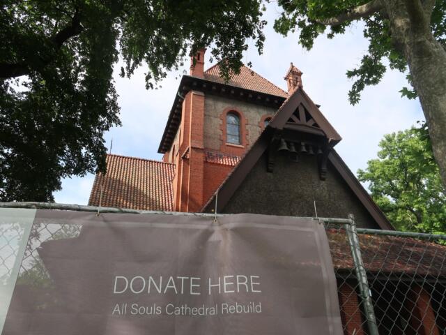 Brick church with red-tiled roof visible through surrounding trees; foreground banner reads “Donate Here” for All Souls Cathedral rebuild.