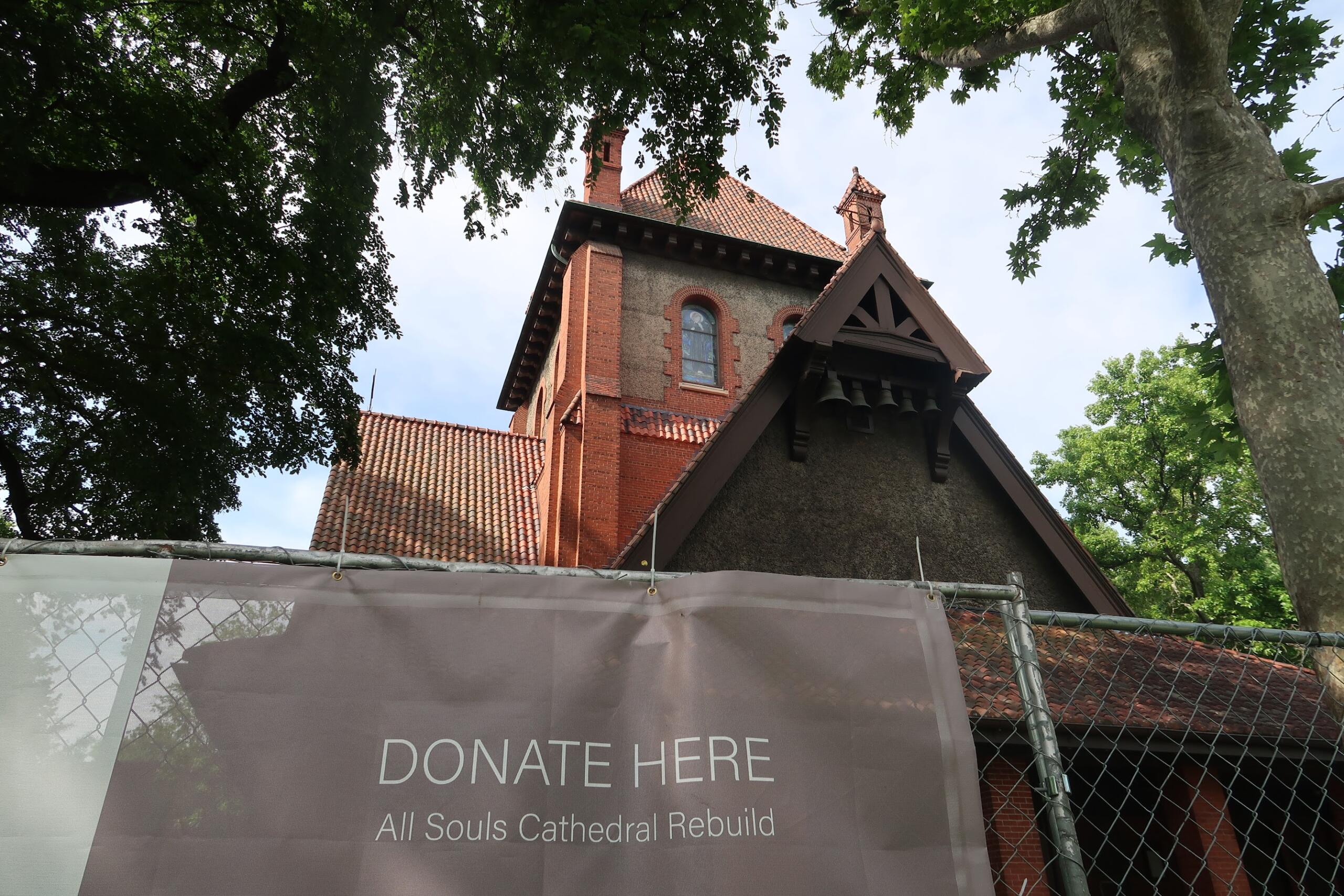 Brick church with red-tiled roof visible through surrounding trees; foreground banner reads “Donate Here” for All Souls Cathedral rebuild.