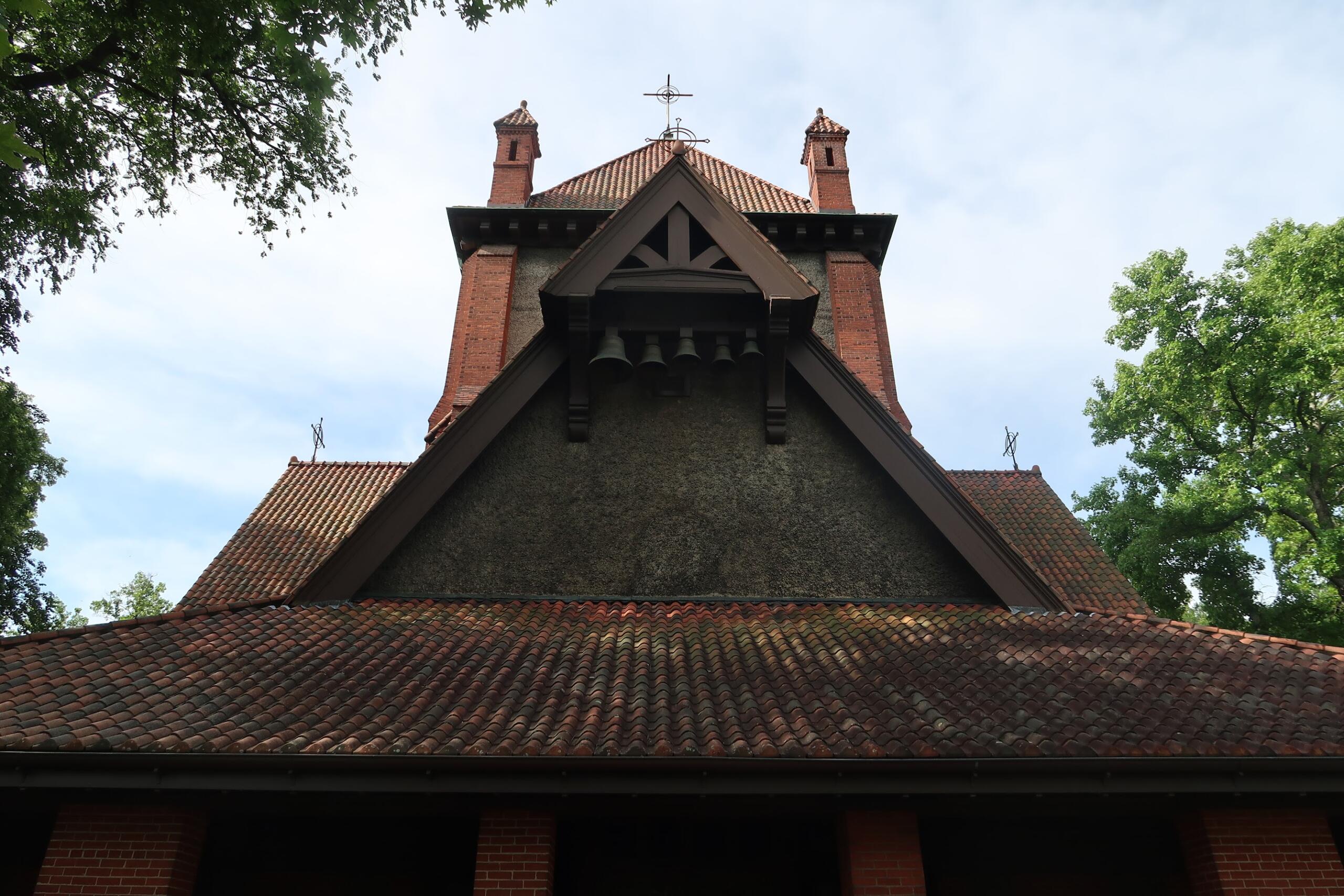 Brick church roof with a triangular gable and hanging bells beneath, flanked by two small brick chimneys, set against a blue sky and surrounding trees