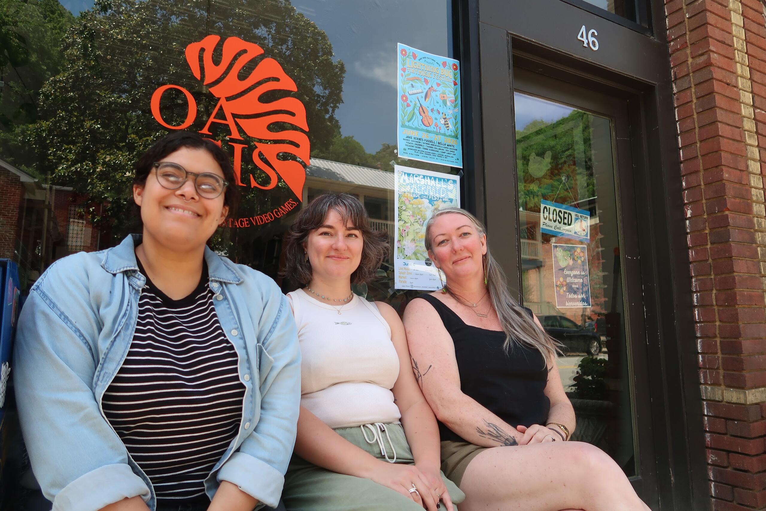 Three smiling women posing for a casual selfie outside a brick storefront with posters in the window and a large orange logo visible on the glass.