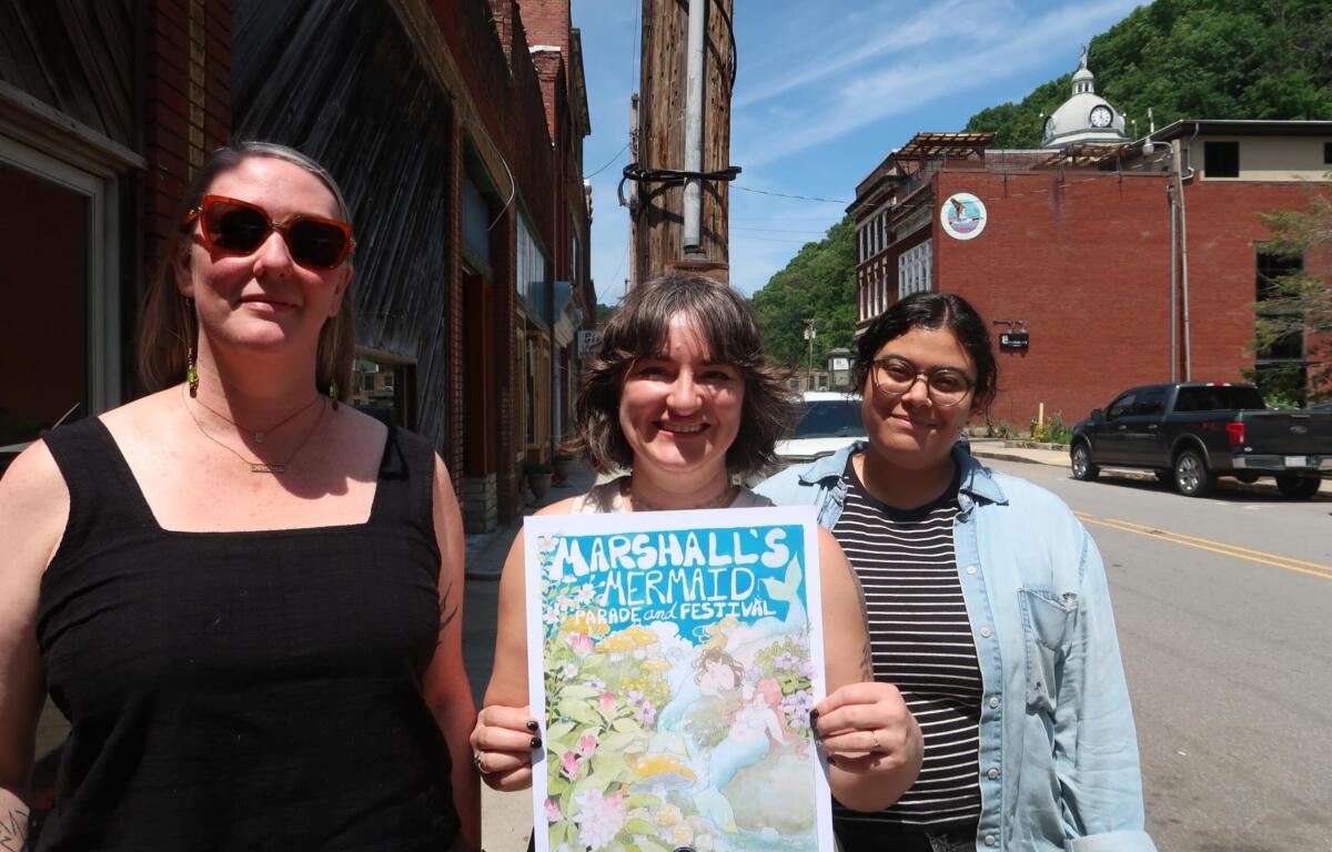 Three smiling women stand on a sunny street, holding a colorful parade poster saying 'Marshall's Mermaid Parade & Festival'.