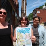 Three smiling women stand on a sunny street, holding a colorful parade poster saying 'Marshall's Mermaid Parade & Festival'.