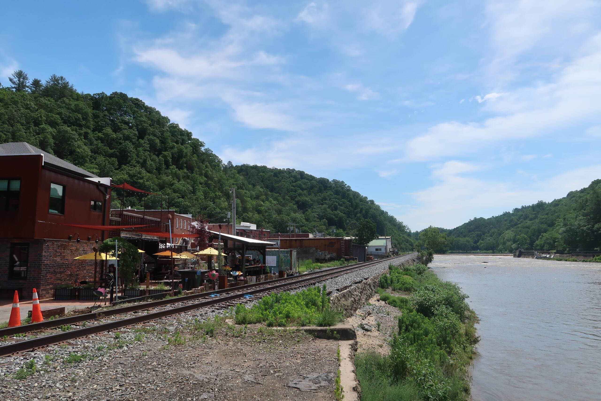 Riverside scene with a railway track, red brick buildings with outdoor seating, and green forested hills under a blue sky.