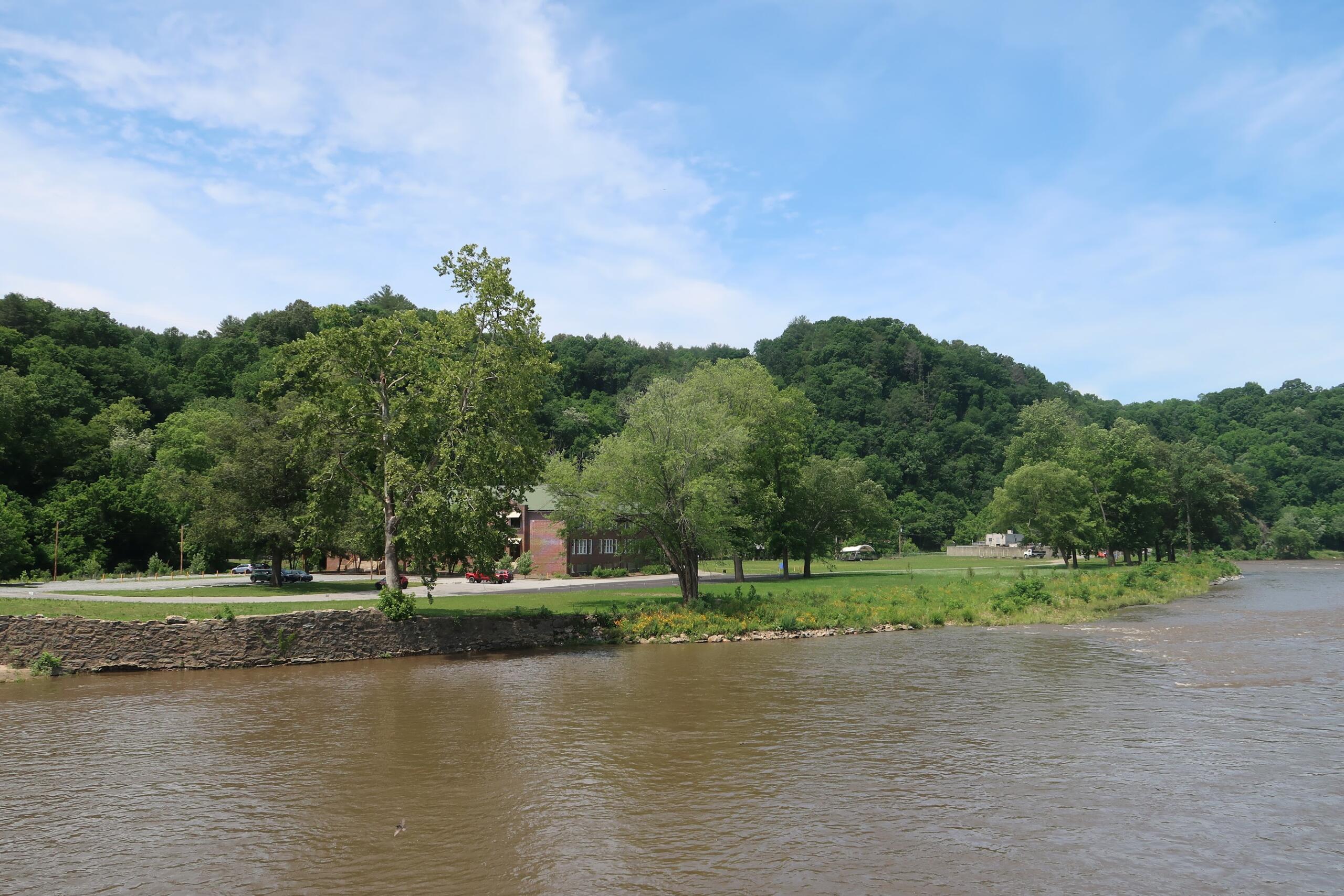 Riverside park with a stone embankment, green grass, and dense trees along a brownish river under a blue sky.