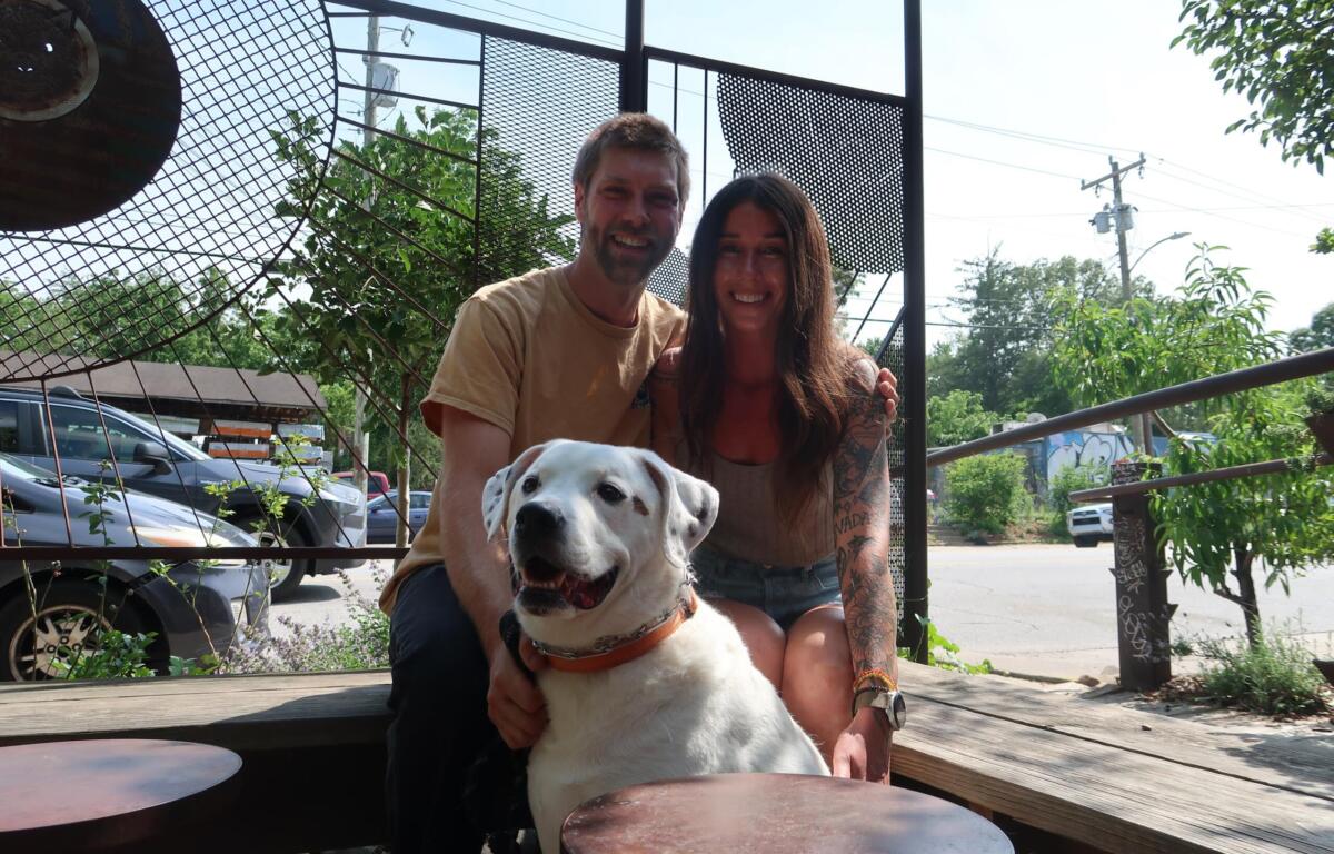 Smiling couple sitting with a white dog on a wooden deck outdoors, with a metal lattice fence and greenery in the background.